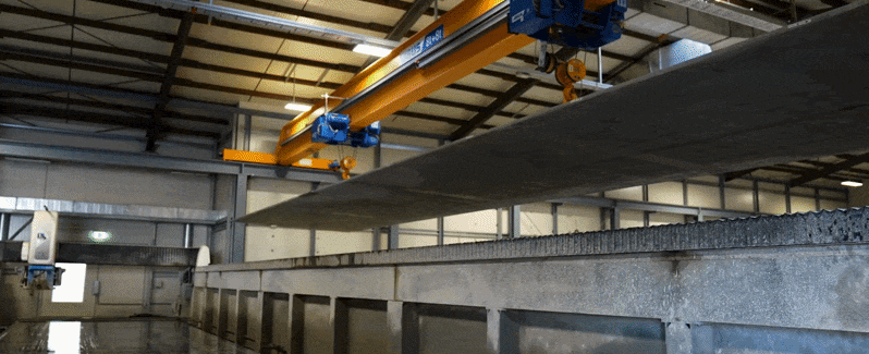Ein Mitarbeiter in Schutzkleidung und Handschuhen kippt eine Probe frischen Carbonbetons aus einem Eimer. Der Beton ist zähflüssig und grau, während der Mitarbeiter den Eimer vorsichtig über einem Behälter hält, um den Beton aufzufangen. Die Szene spielt in einer industriellen Umgebung, möglicherweise in einer Produktionshalle oder einem Labor, mit unscharfem Hintergrund, der die Aufmerksamkeit auf die Aktion des Mitarbeiters lenkt.